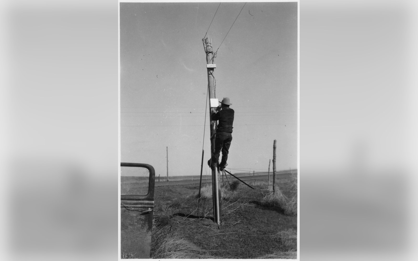 A lineman working at the top of a wooden pole against a flat sky